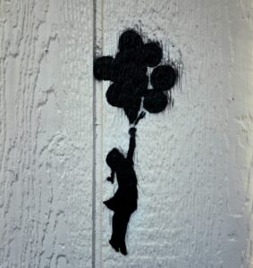 Stencil-style street art of a young girl reaching upward toward a cluster of balloon shapes, painted in black on a pale wooden surface.