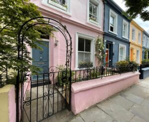 Pastel-coloured terraced houses in Notting Hill, with pink, blue and yellow façades, wrought-iron railings and leafy trees framing the street.