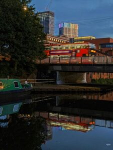 A green narrowboat on the Grand Union Canal in London with a red double-decker bus crossing a bridge in the background.
