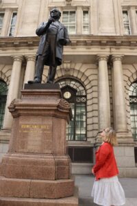 Guide Hazel standing beside the statue of Rowland Hill outside the Royal Exchange in the City of London, looking up at the Victorian postal reformer